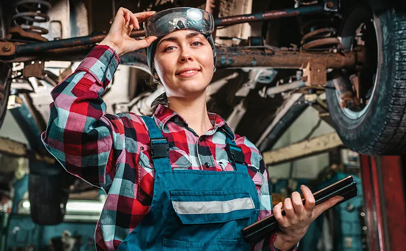 Jeune femme en tenue de travail et lunettes de protection dans un atelier de mécanique, tenant une tablette numérique, représentant l’alternance en formation professionnelle.