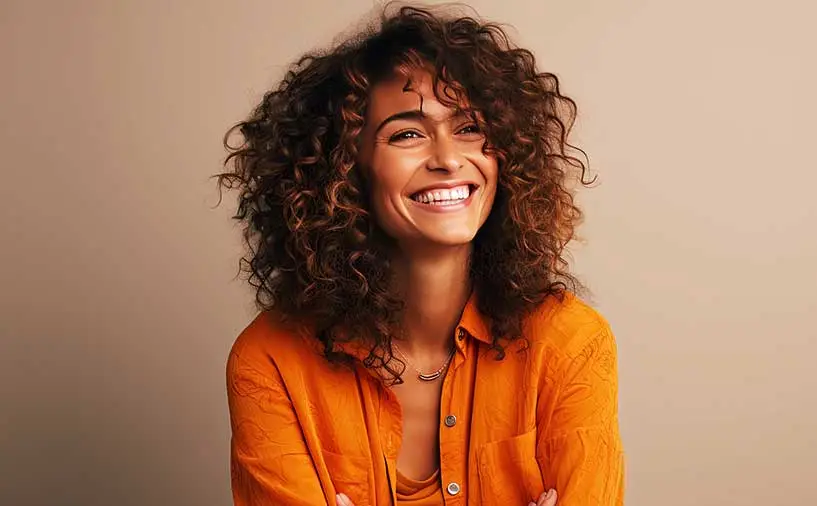 Portrait d’une jeune femme souriante, aux cheveux bouclés, portant une chemise orange, posant devant un fond uni beige. L’image évoque la confiance, la détermination et l’optimisme.