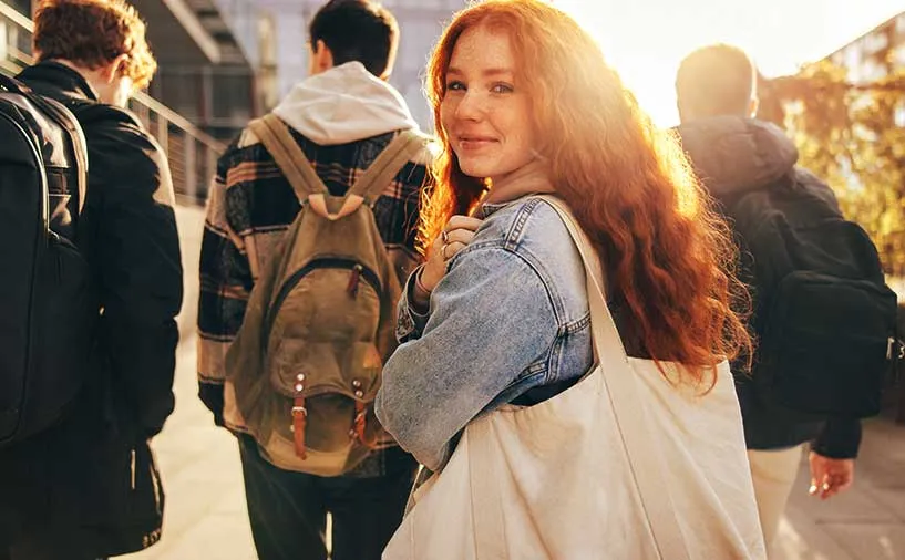 Une jeune femme rousse souriante, entourée d'autres jeunes, marche en extérieur avec un sac en toile sur l’épaule, illustrant l’accompagnement des jeunes en mission locale.