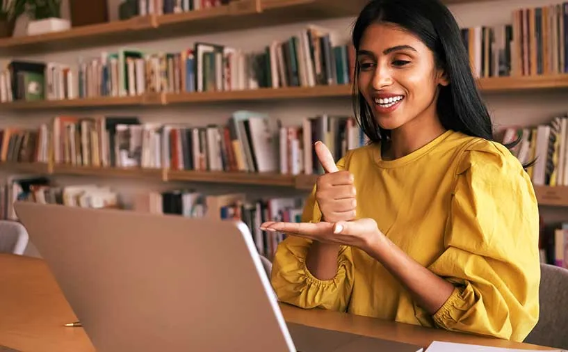 Femme souriante en visioconférence utilisant la langue des signes française, devant un ordinateur, dans une bibliothèque.