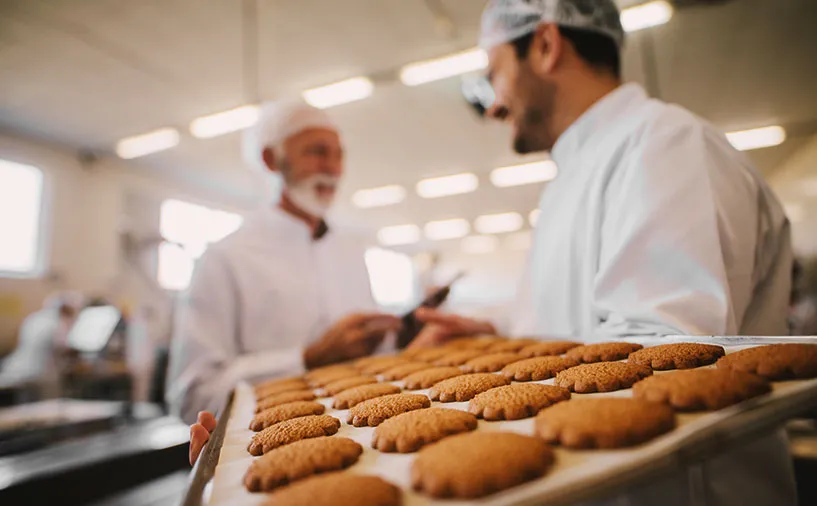 Deux professionnels en tenue de laboratoire discutant dans une biscuiterie, avec un plateau de biscuits en premier plan.